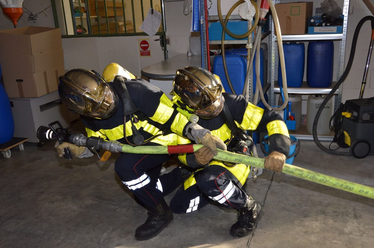 Two firefighters in full PPE pulling a fire hose through a warehouse.